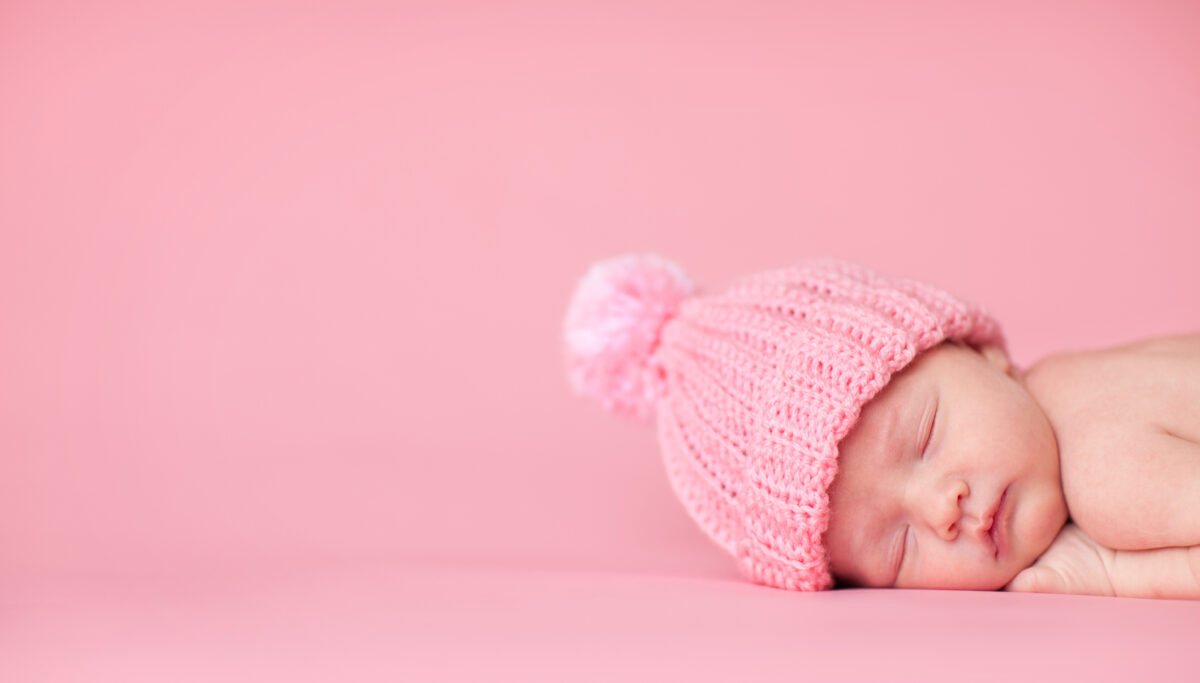 Color photo of a beautiful newborn baby girl wearing a knit hat while sleeping peacefully on a pink background.