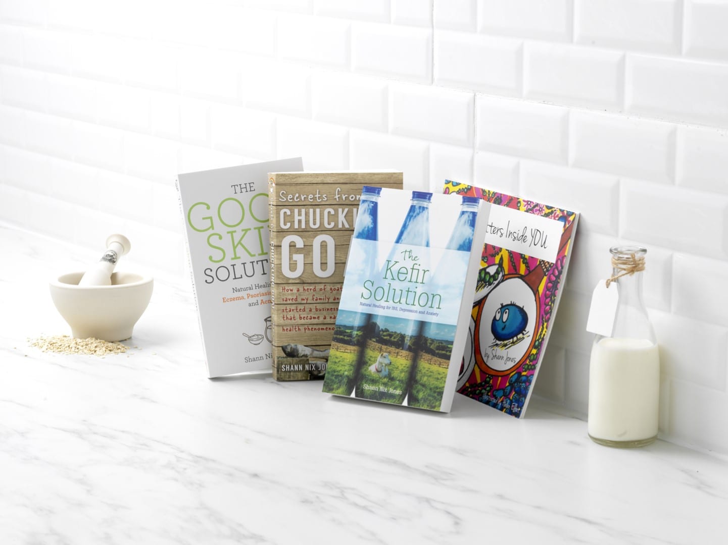 Several Chuckling Goat books on a kitchen countertop next to a bottle of kefir and a pestle and mortar