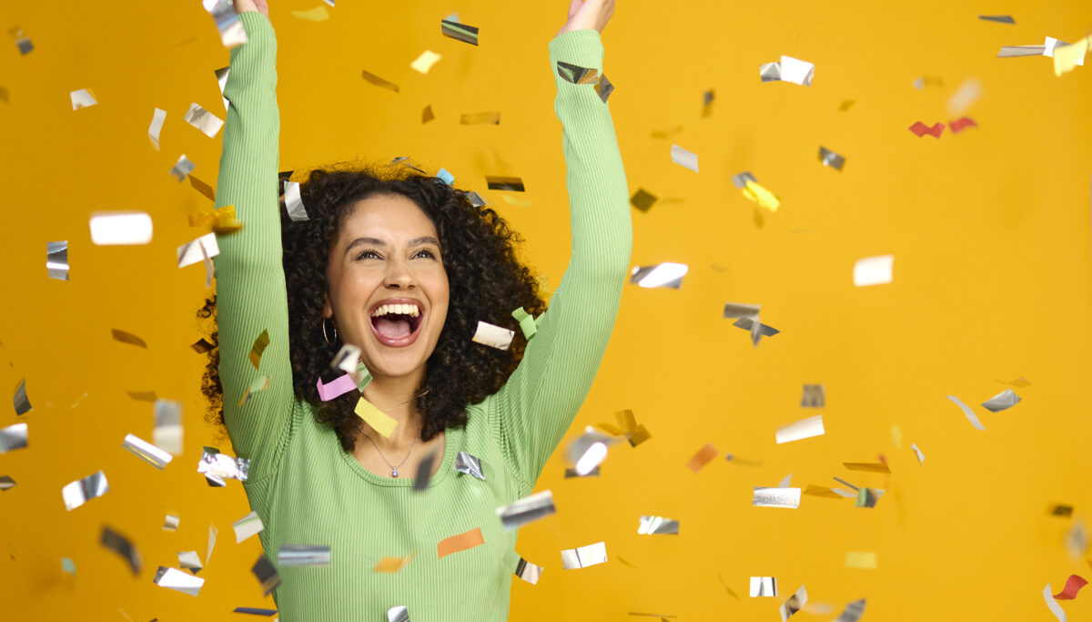 Studio Shot Of Excited Woman Celebrating Big Win Showered In Tinsel Confetti On Yellow Background