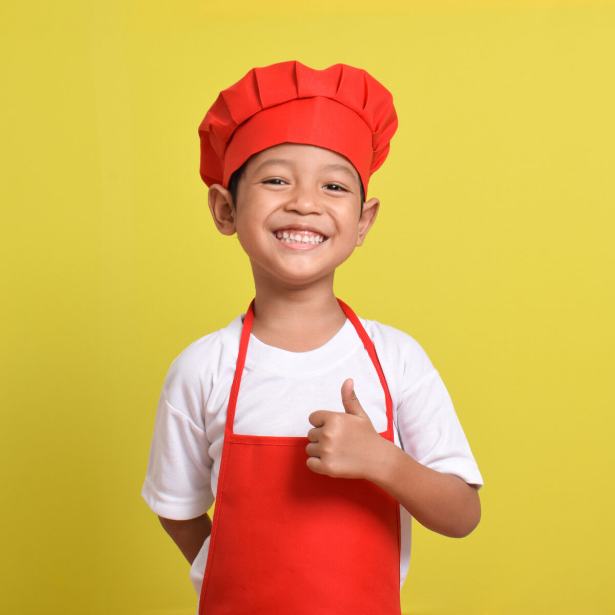 Cute little chef showing thumbs up isolated on yellow background, wearing red apron and hat.
