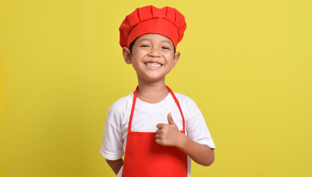 Cute little chef showing thumbs up isolated on yellow background, wearing red apron and hat.