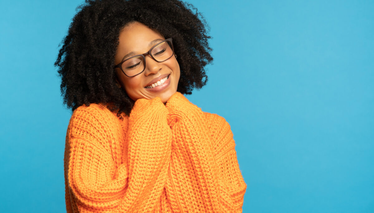 Stylish happy dark skinned millennial woman with curly hair wear orange knitted jumper, holds hands under chin, copy space for advertising, isolated on studio blue background. Winter warm clothes.