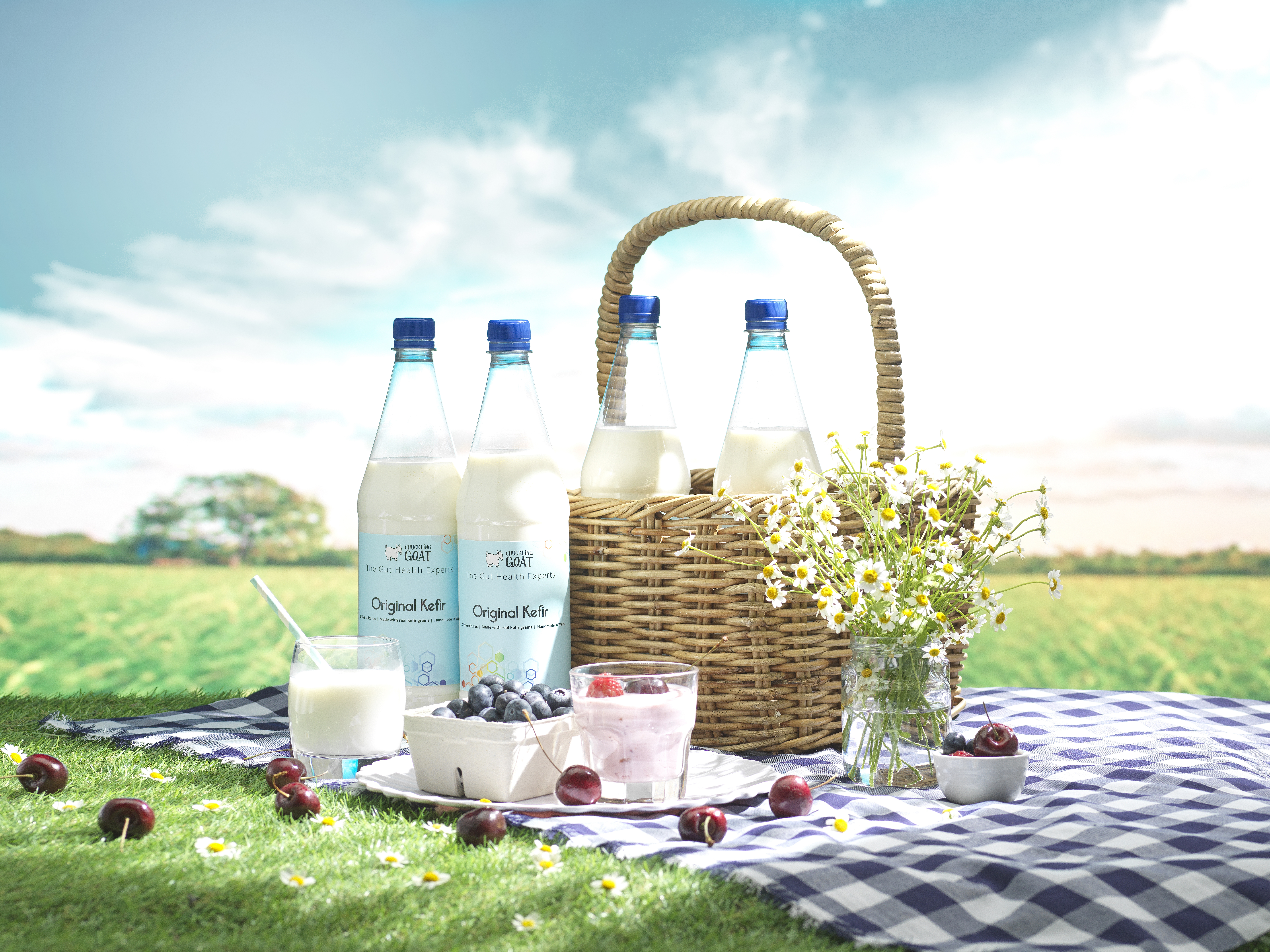 Image of Chuckling Goat Kefir bottles on a picnic blanket with picnic basket. Blue skies and green grass as the background.