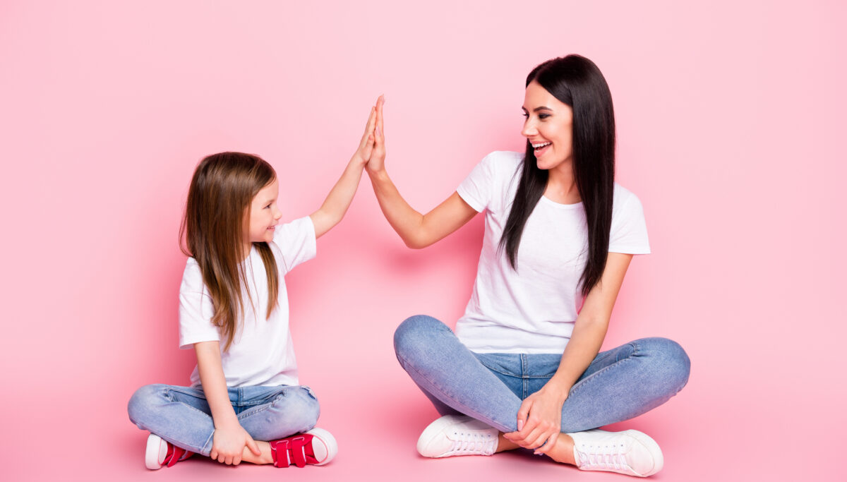 Full body photo of young mom small daughter best friends, sit floor crossed legs clap arms cool team wear casual white t-shirts jeans shoes isolated pastel pink colour background