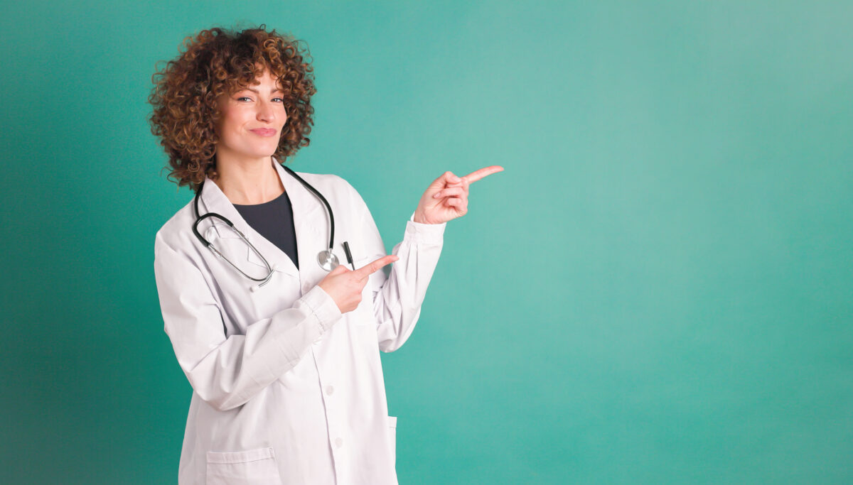 Smiling young curly haired female looking at camera pointing index fingers at empty green background