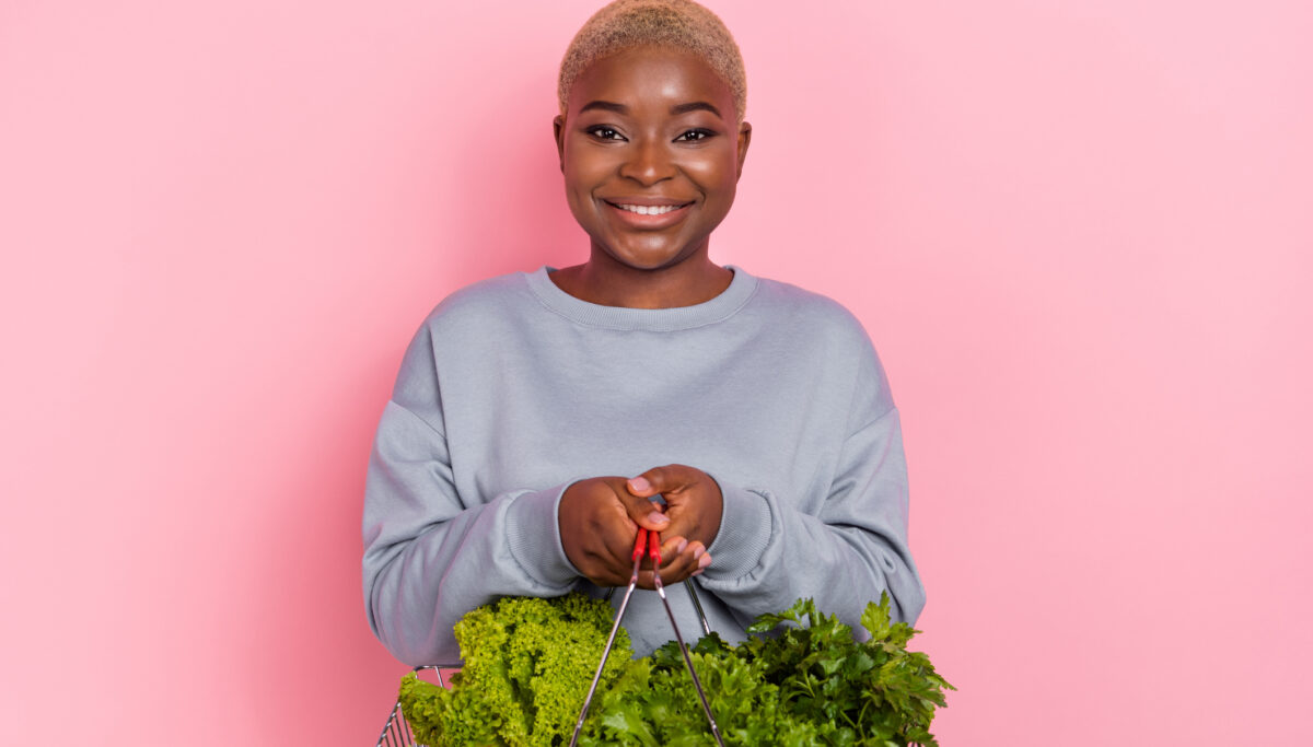 Woman smiling holding a shopping basket full of plant foods on a pink background.