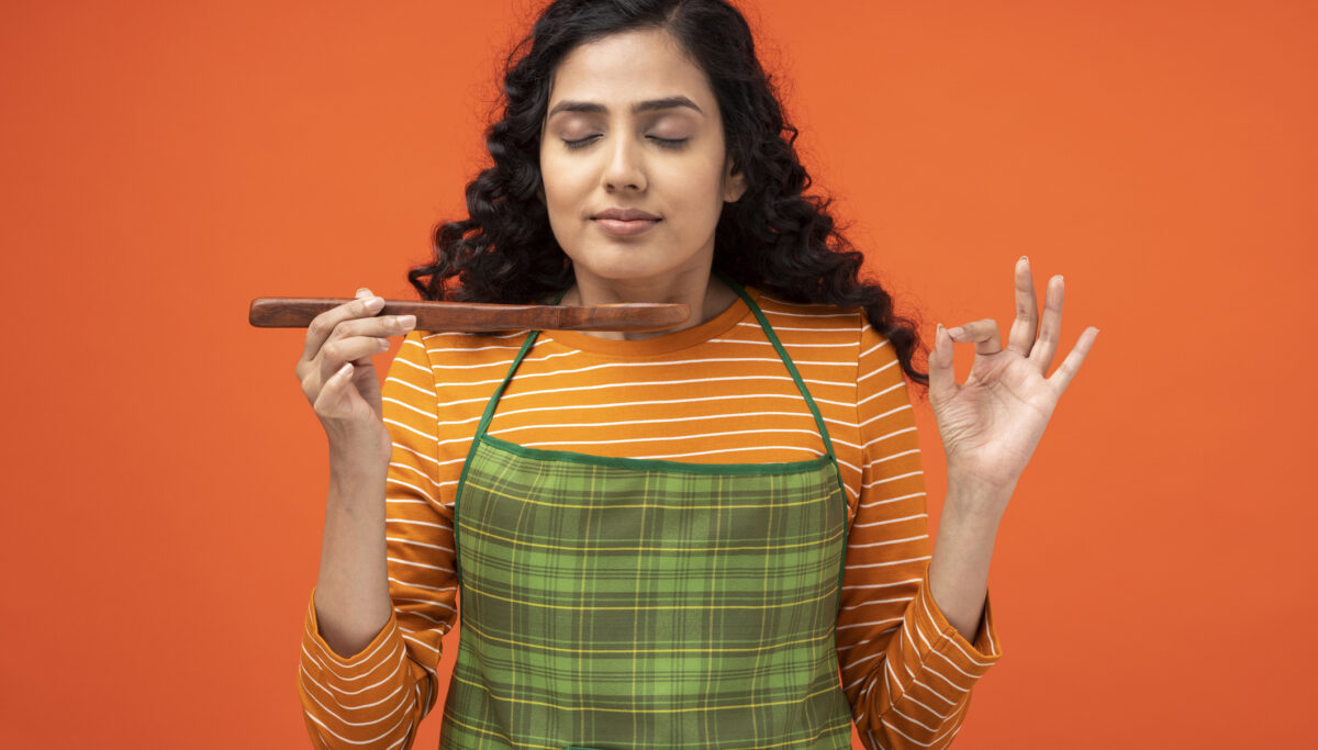Woman cooking, holding a wooden spoon and wearing an apron, on an orange coloured background.