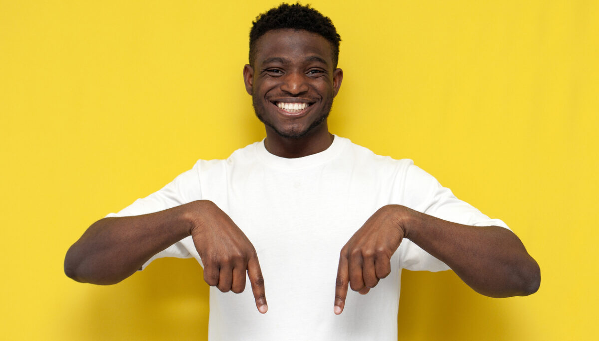 Joyful African American man in white t-shirt shows his hands pointing down on yellow isolated background.