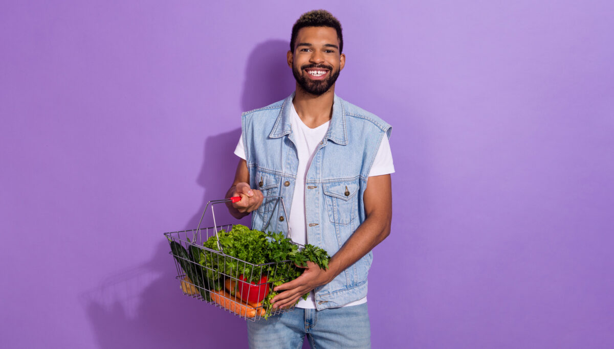 Man smiling holding a basket of natural, wholefoods on a purple background.