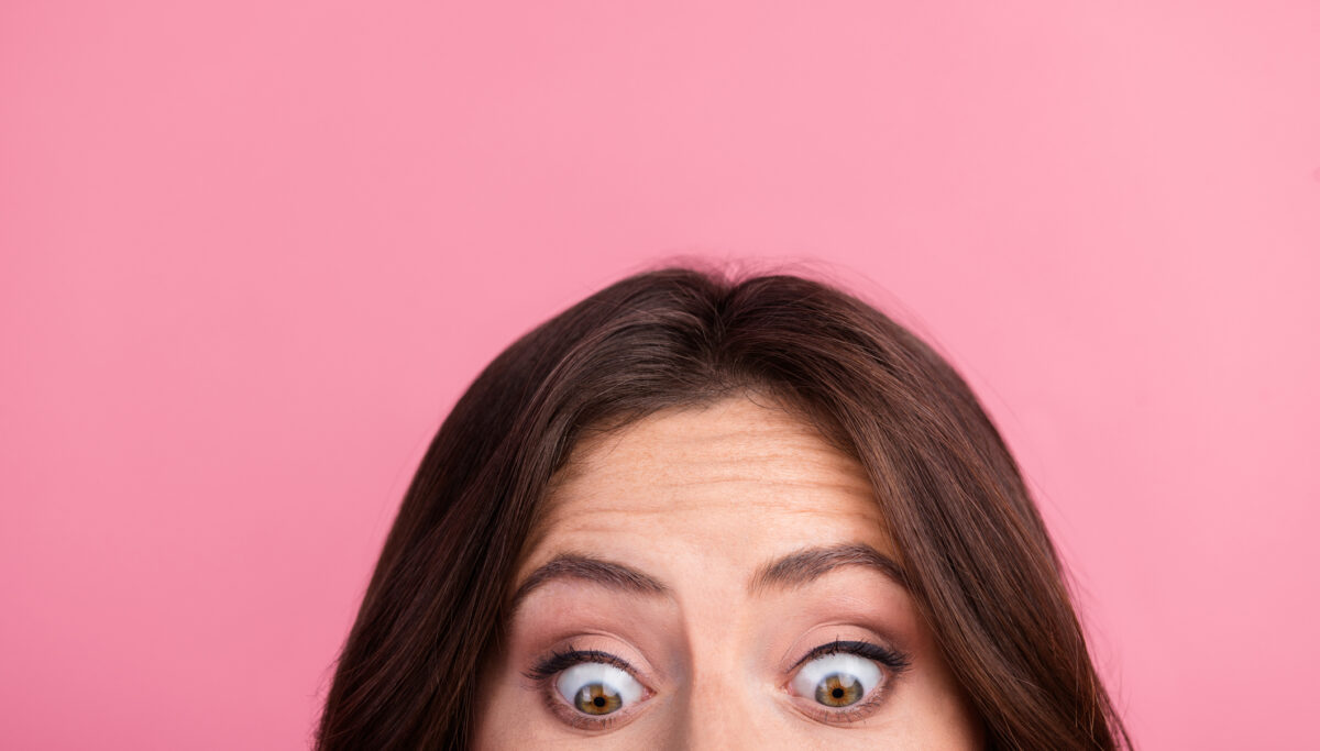Close-up of a young woman's surprised face against a pink background, highlighting her expressive eyes and raised eyebrows, conveying curiosity and wonder.