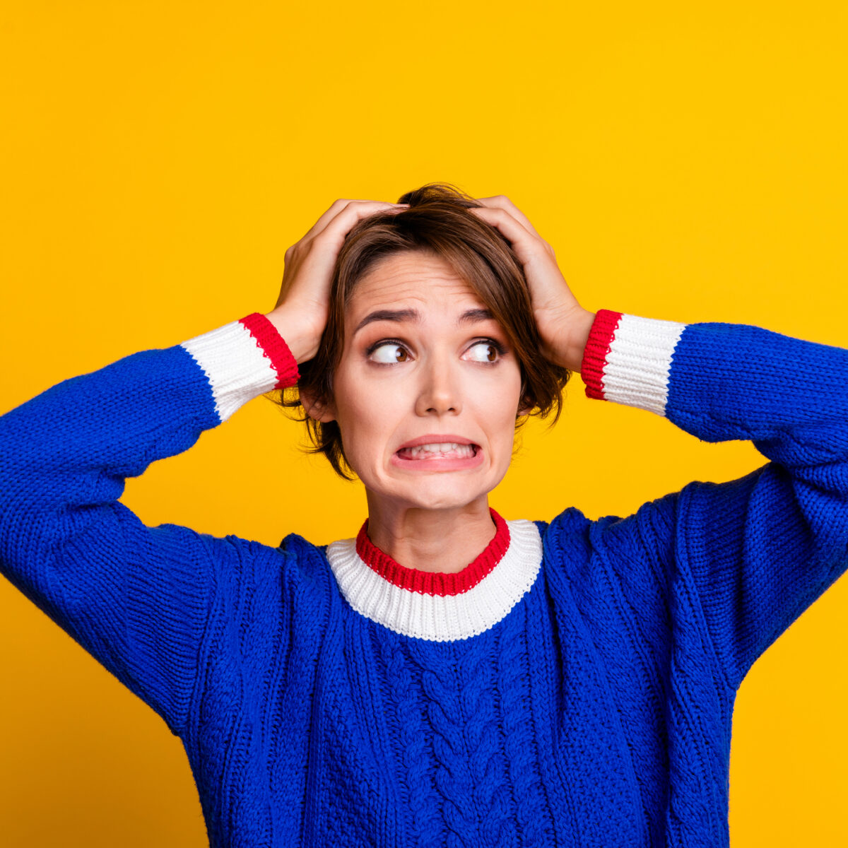 Woman wearing a blue jumper with hands on her head, looking unsure, on a yellow background.