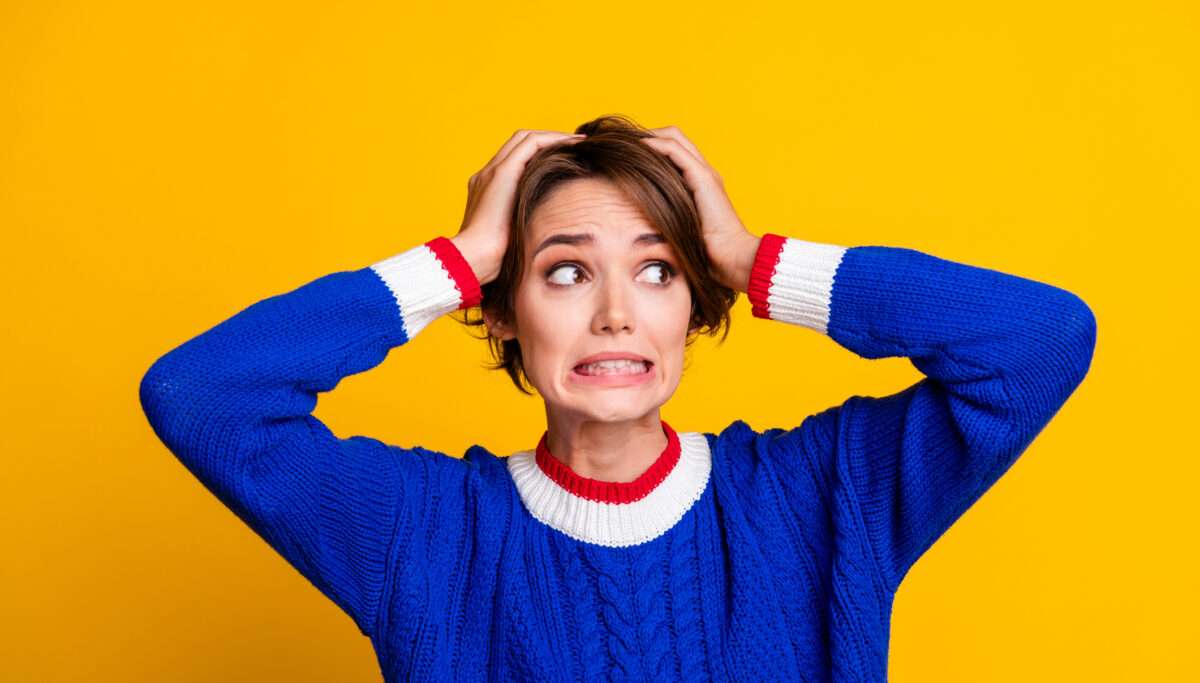 Woman wearing a blue jumper with hands on her head, looking unsure, on a yellow background.
