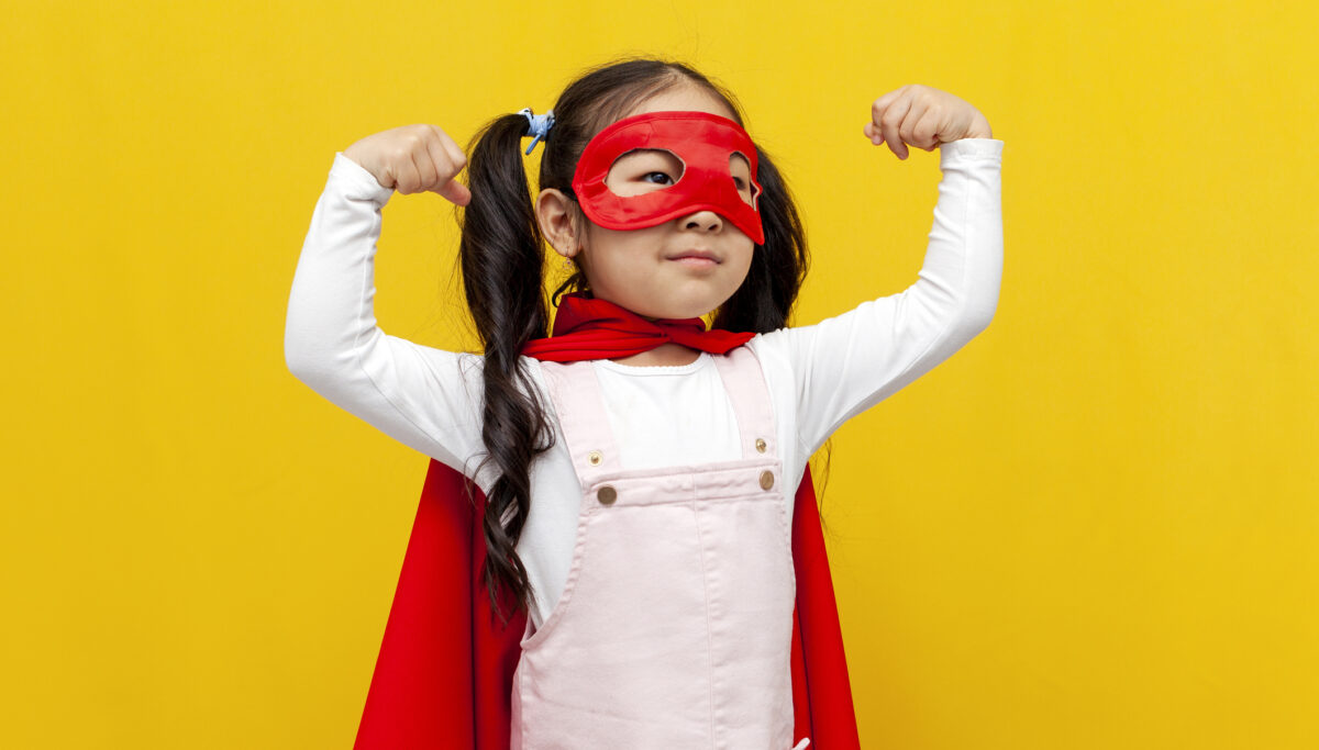 little girl in superman costume and mask showing biceps and power gesture, superhero cape smiling over yellow isolated background