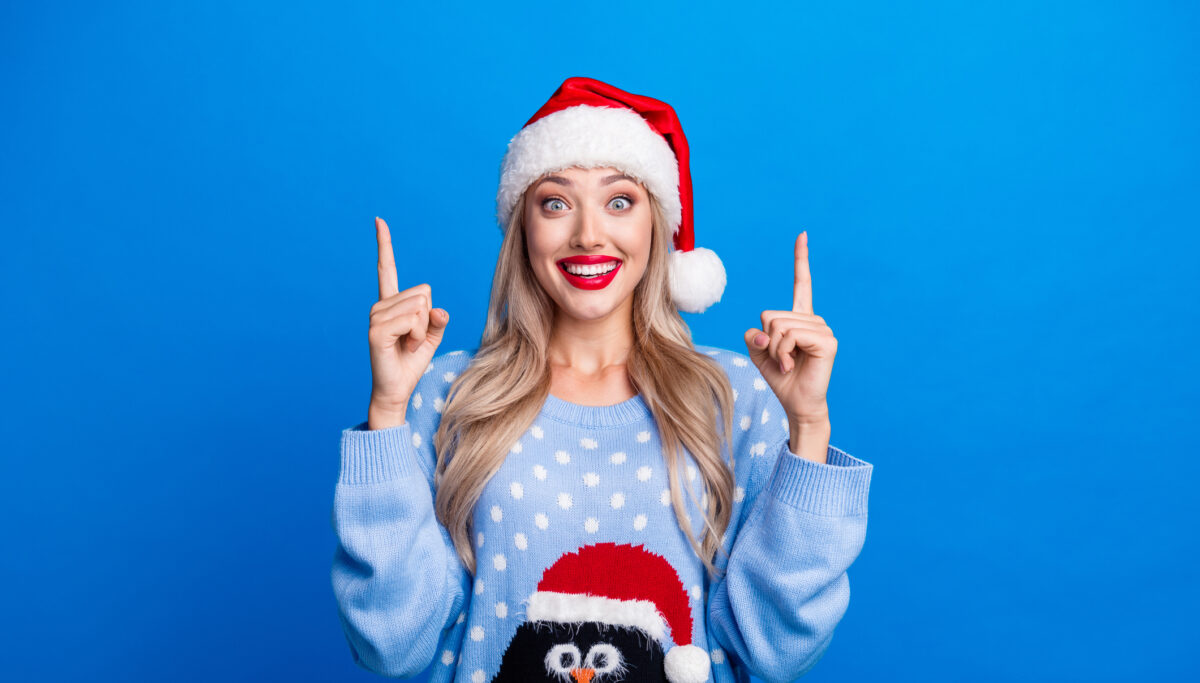 Photo portrait of blonde young girl excited finger point up dressed penguin sweater x-mas hat isolated on blue color background.