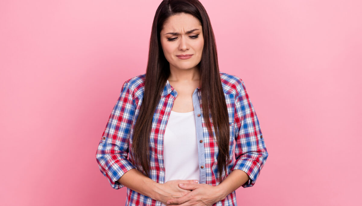 Woman holding her stomach in pain on a pink coloured background.