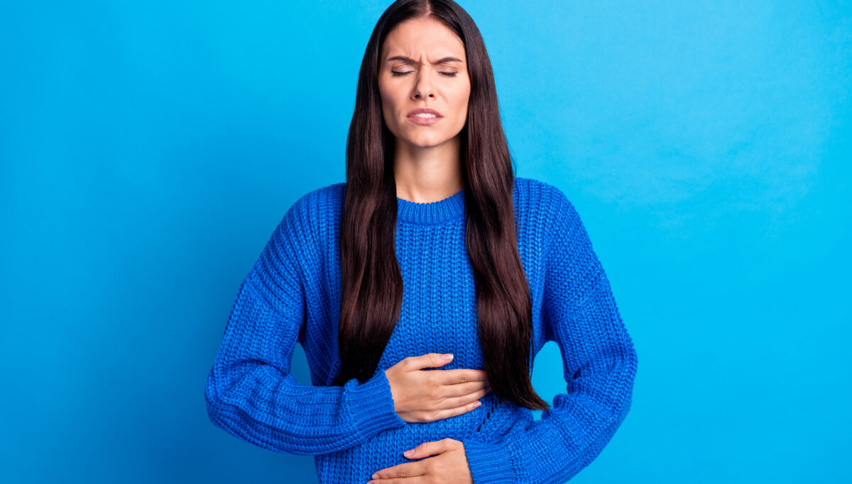 Woman holding stomach with both hands and visibly uncomfortable and in pain, on a blue background.