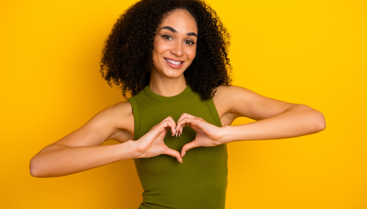 Smiling young woman with curly black hair holds hands in a heart shape. Yellow background and green top emphasize her cheerful personality and vibrant energy.