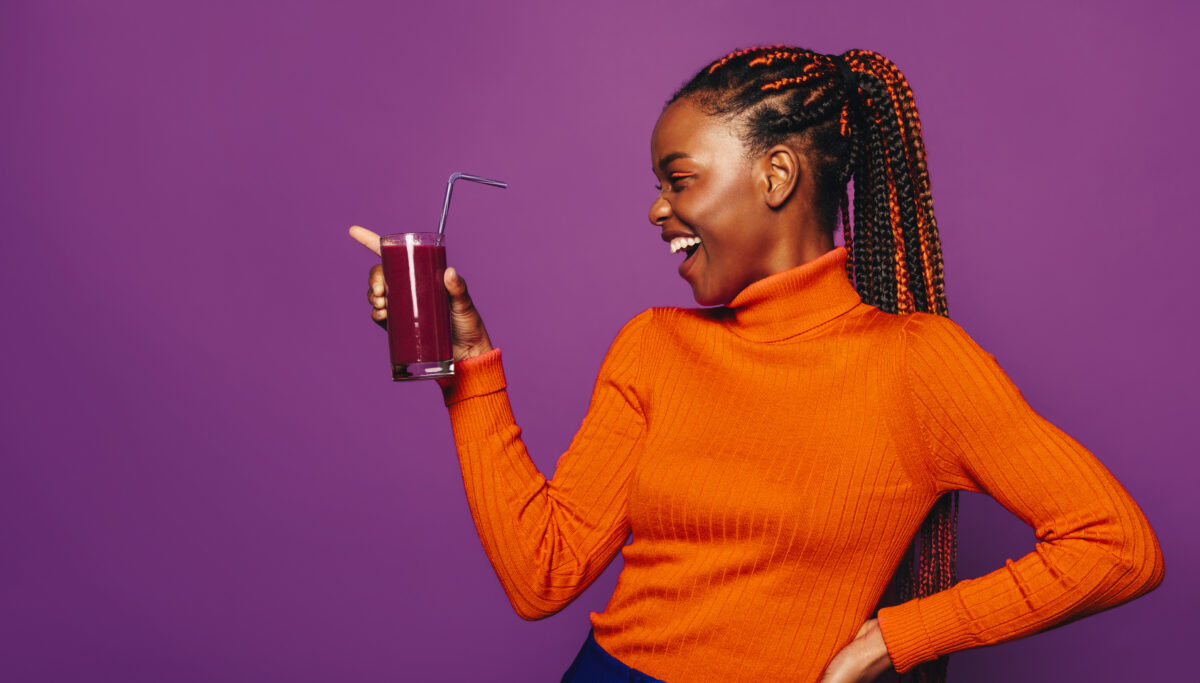 Cheerful young woman with stylish two-tone braids stands in a vibrant purple studio. She holds a cup with a straw, enjoying a fresh and healthy smoothie. Her casual clothing and happy smile reflect a fun-loving lifestyle.