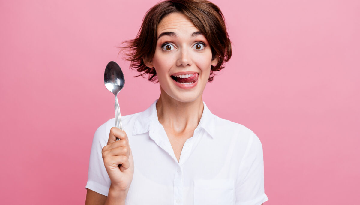 Portrait of young girl lick teeth eat delicious dessert holding silver spoon excited isolated on pink color background.