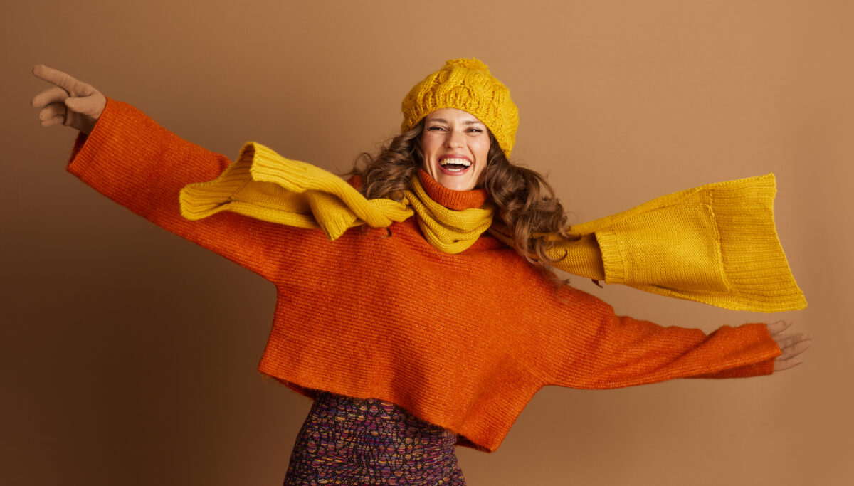 A joyful young woman with curly brown hair wears a vibrant orange sweater, yellow knit scarf, and hat. Her arms are outstretched in a dynamic, celebratory pose against a warm brown background.
