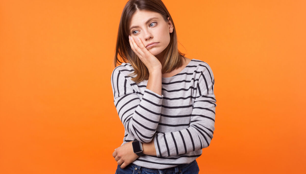 Portrait of bored young woman with brown hair in long sleeve shirt standing with face in hand, thinking sadly, looking depressed and disinterested. indoor studio shot isolated on orange background