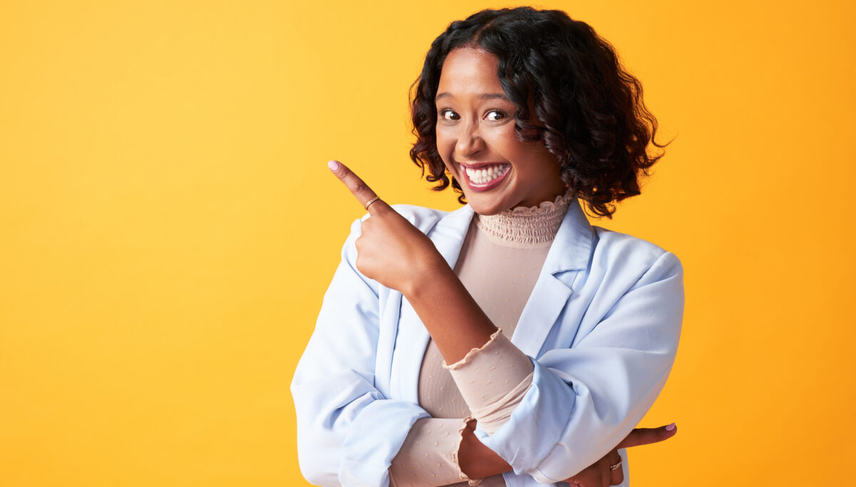Excited, science and trendy woman pointing her finger, showing direction or raising her hand standing against a yellow background. Trendy, happy and cheerful female making hand gesture