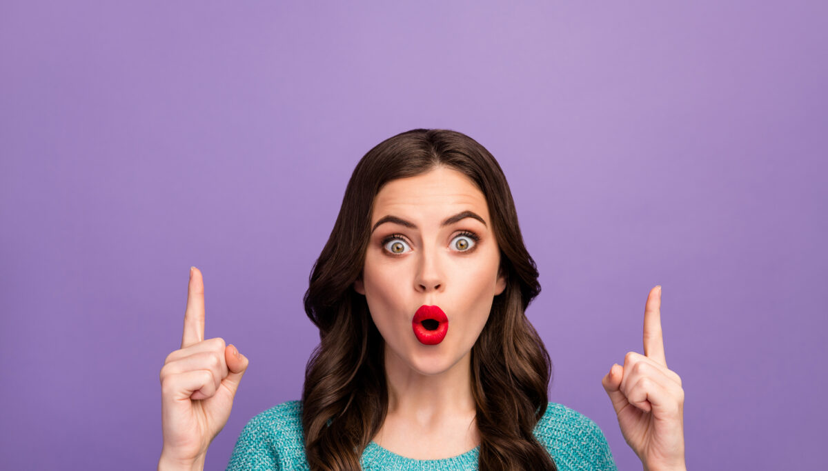 Surprised woman wearing red lipstick, pointing upwards, purple solid background
