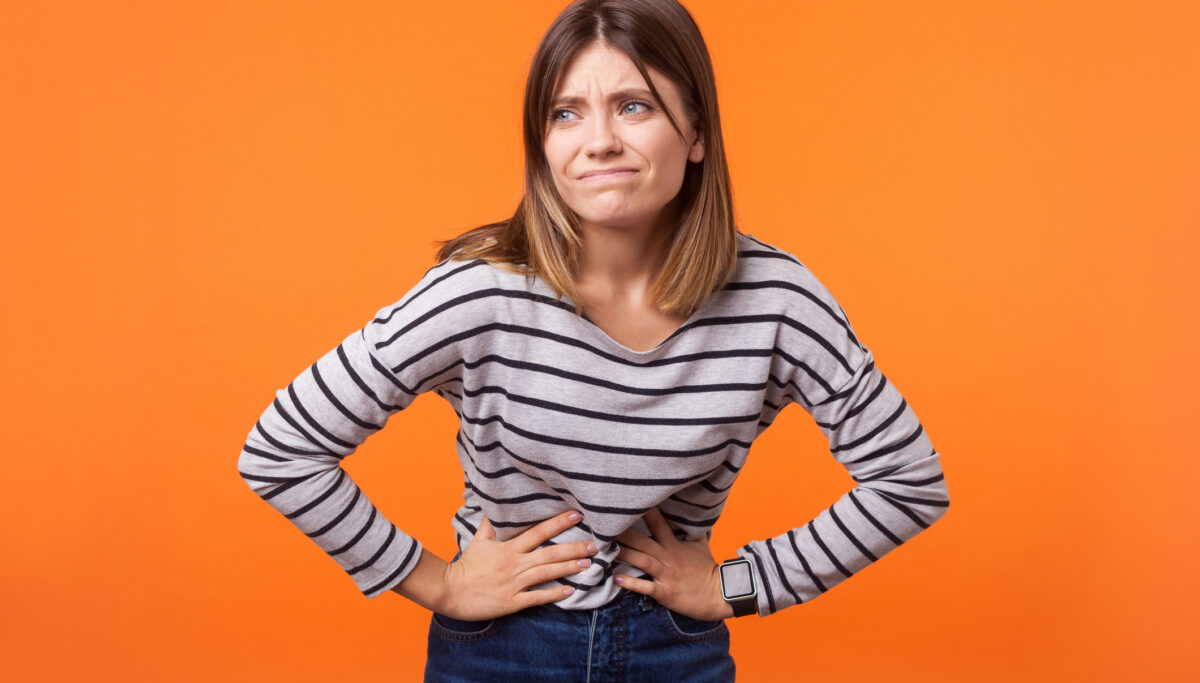 Portrait of woman with brown hair, holding tummy, uncomfortable face, stomach cramps, on isolated orange background