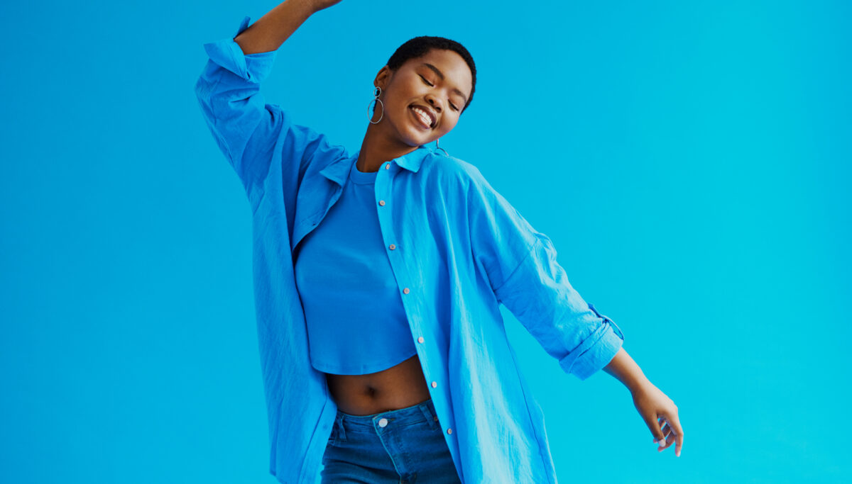 Young black woman wearing a jean, blue shirt and blue top whilst dancing and smiling shot against a blue background with copy space stock photo
