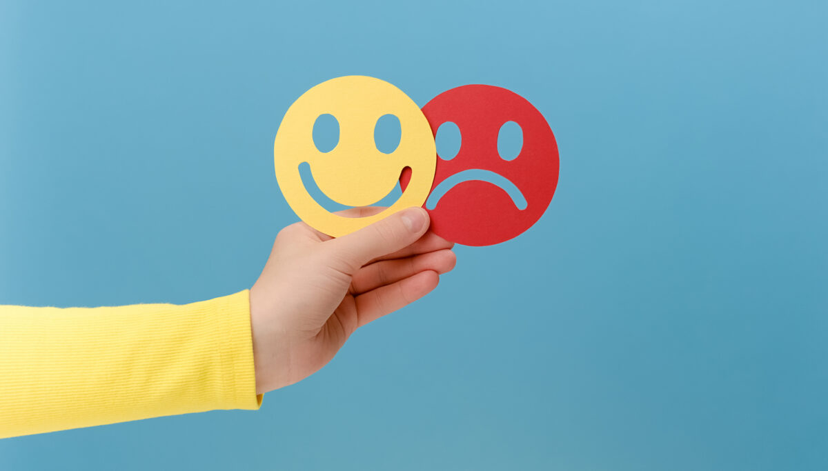 Hand holding happy and sad cut-out paper faces on isolated blue background