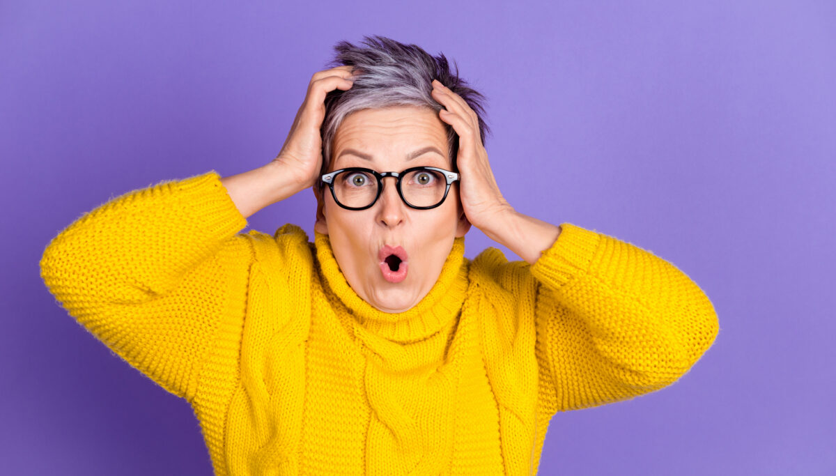 A mature woman in a yellow sweater and glasses, holding head in shock or surprise. Vivid purple background.