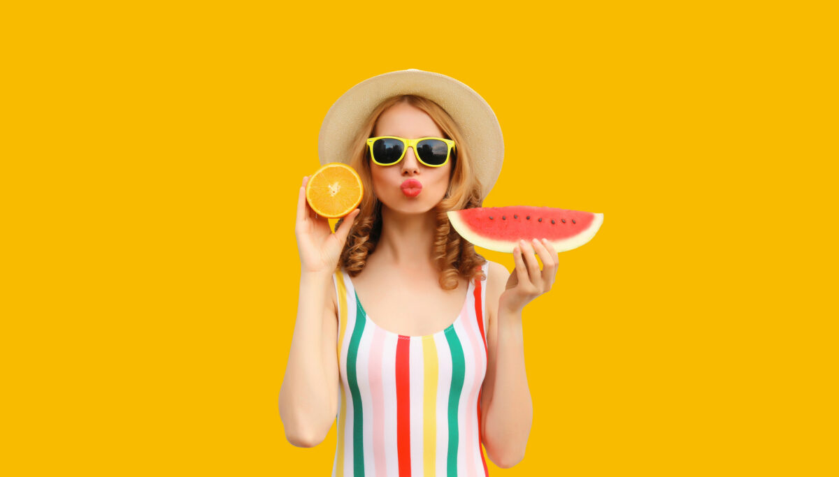 Summer portrait of happy young woman posing with fresh juicy fruits, slice of watermelon and orange wearing straw hat, sunglasses on colorful yellow background