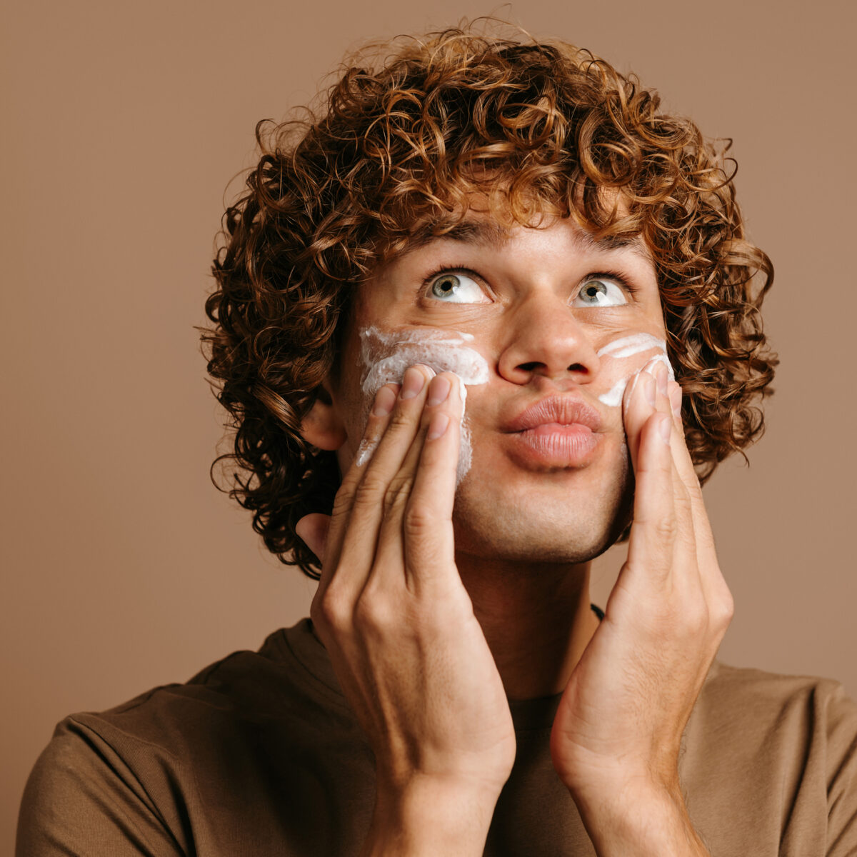Handsome man enjoying beauty treatment while applying cream on face and standing on brown background