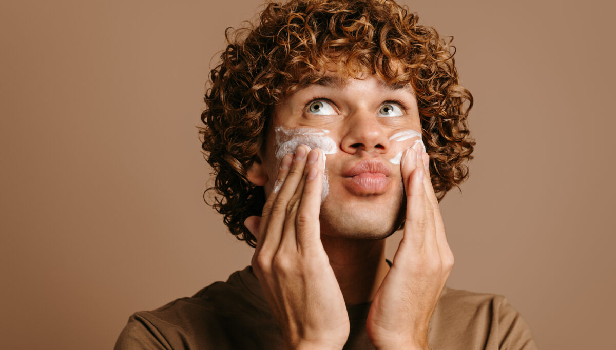 Handsome man enjoying beauty treatment while applying cream on face and standing on brown background