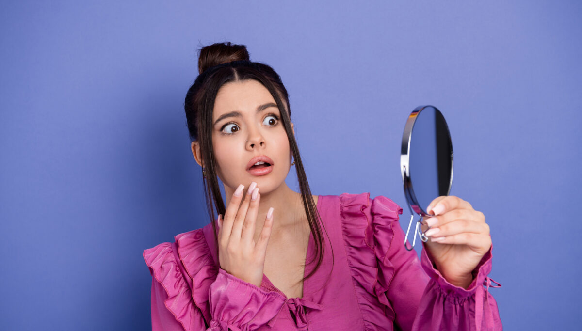 A young brunette woman in a trendy pink outfit looks shocked while holding a handheld mirror, set against a violet background.