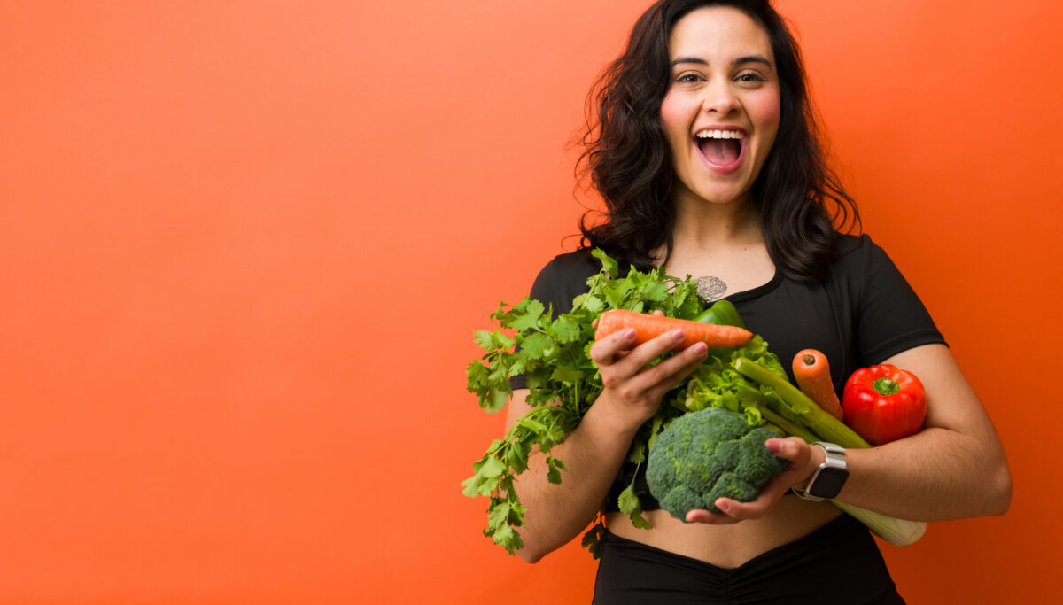 Hispanic woman holding a variety of fresh vegetables. Showing excitement for healthy eating, fitness, and vitality on an orange studio background