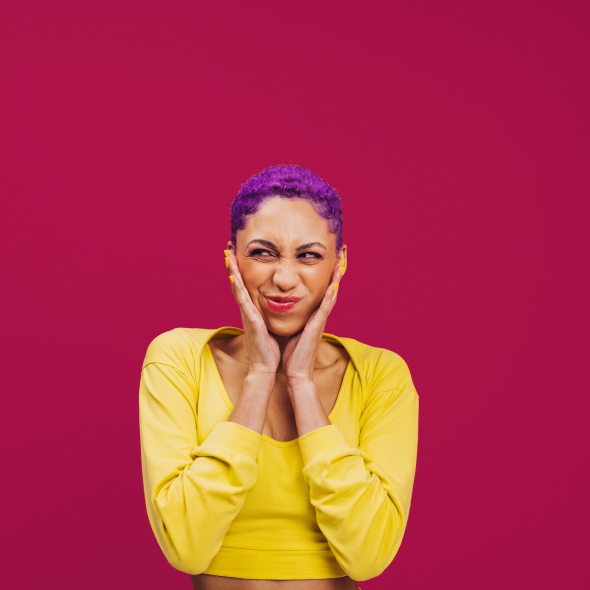 Being quirky in the studio. Playful young woman making a funny expression while standing against a pink background. Fashionable young woman wearing makeup with purple hair in a studio.