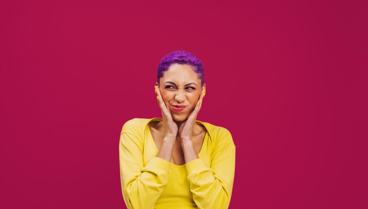 Being quirky in the studio. Playful young woman making a funny expression while standing against a pink background. Fashionable young woman wearing makeup with purple hair in a studio.