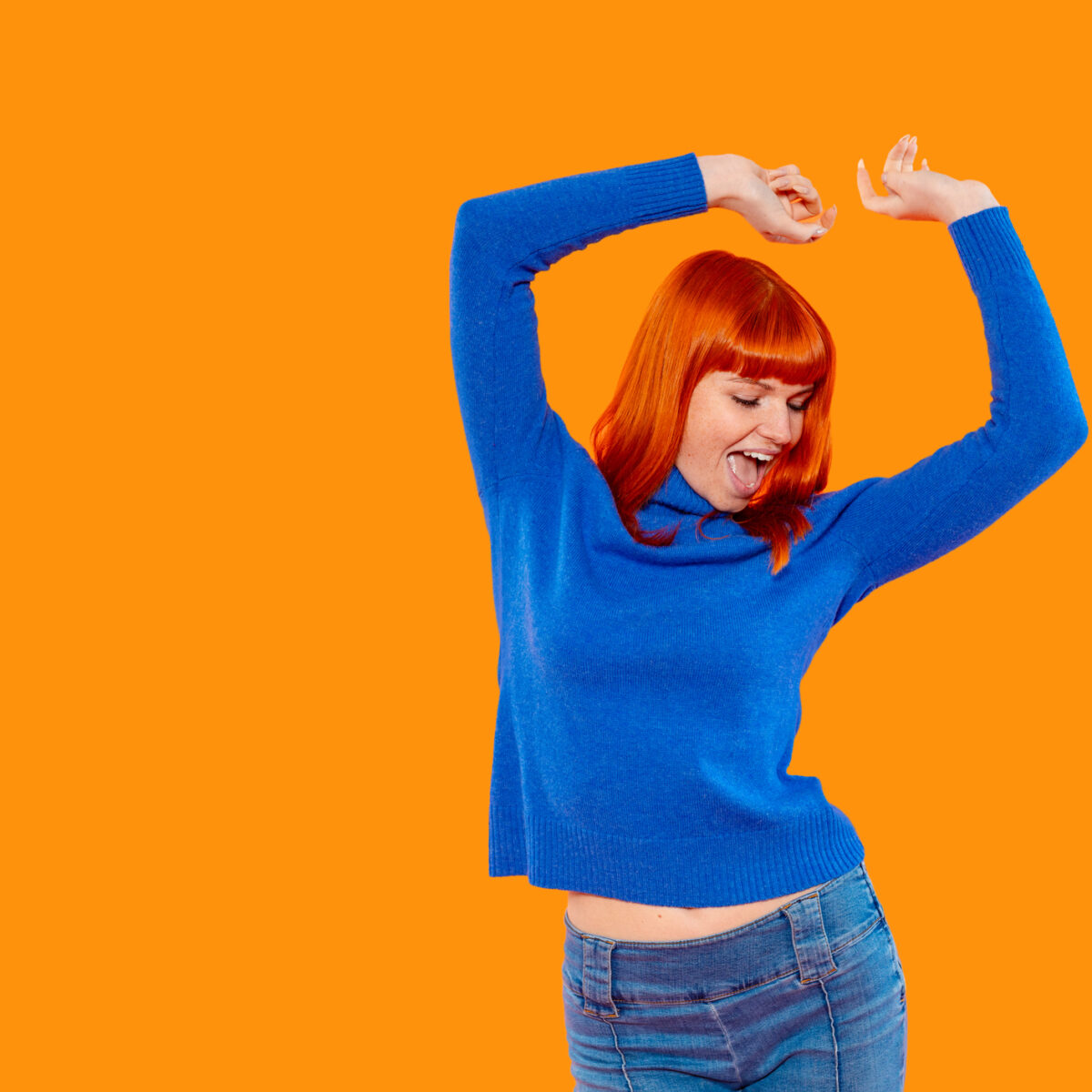 A joyful woman with bright red hair dances enthusiastically in a cozy indoor setting, wearing a blue sweater and showing a carefree attitude against an orange backdrop.