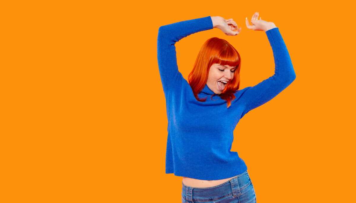 A joyful woman with bright red hair dances enthusiastically in a cozy indoor setting, wearing a blue sweater and showing a carefree attitude against an orange backdrop.