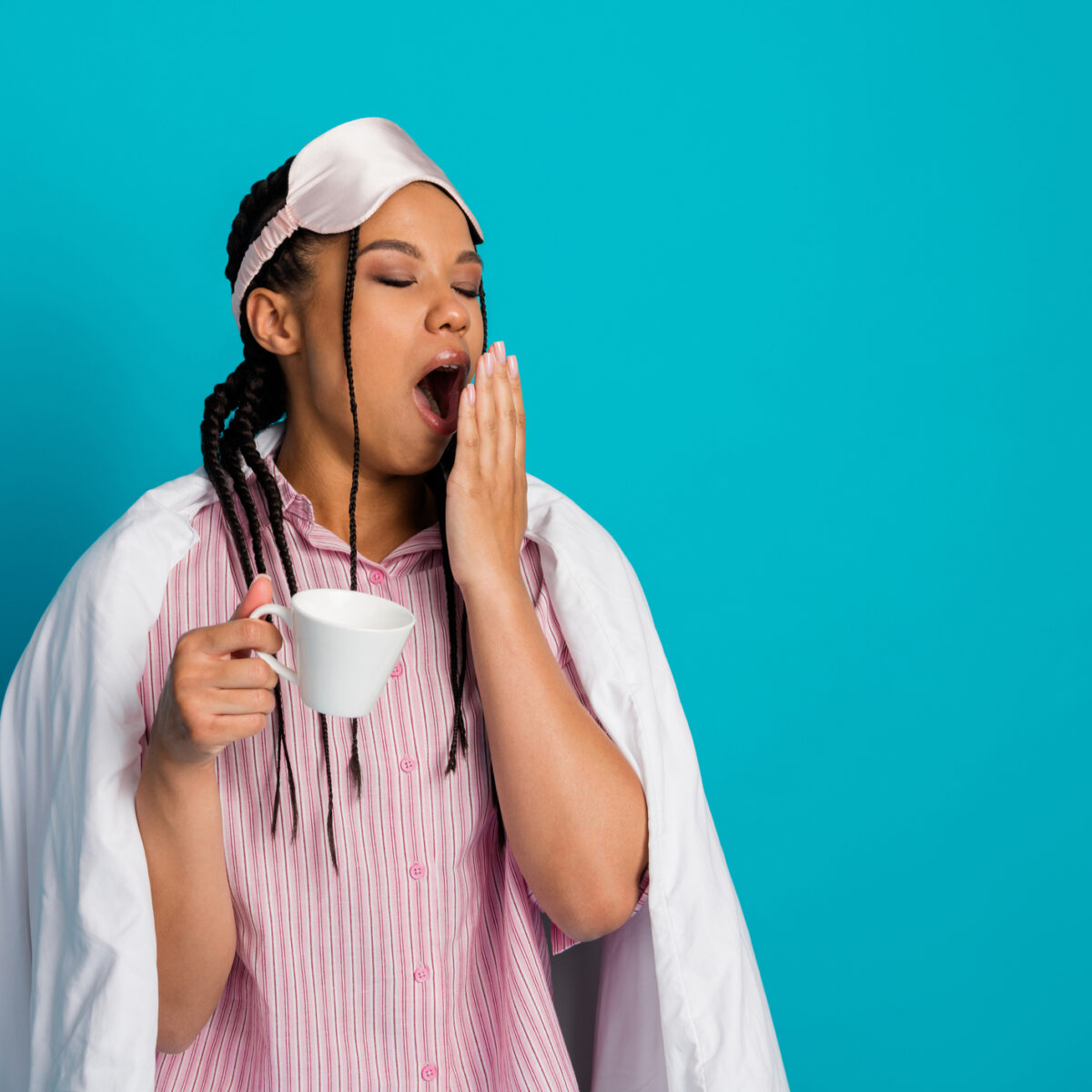Always tired. A young woman wearing pink sleepwear, yawning while holding a cup, against a vibrant blue background