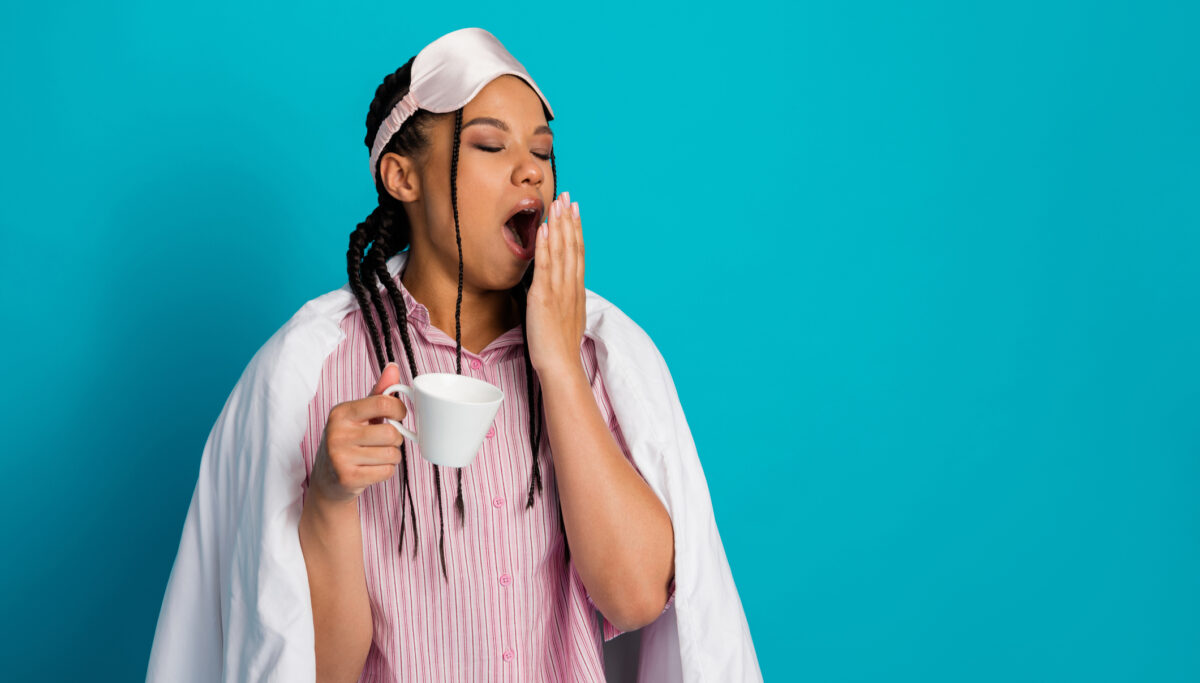 Always tired. A young woman wearing pink sleepwear, yawning while holding a cup, against a vibrant blue background